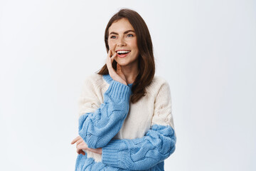 Portrait of young woman staring left at copy space and smiling amused, watching something interesting aside, standing in sweater against white background