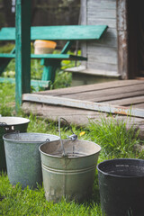 Several old buckets are standing on the grass in sunny weather. Collecting rainwater in buckets in the village. Buckets are in one line. Old wooden house and bench in the background. Toning