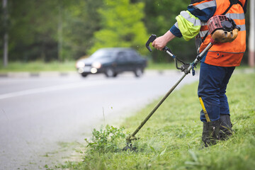 A worker in special reflective clothing with a gasoline mower in his hand. A man with a trimmer mows grass with dandelions on the side of the road
