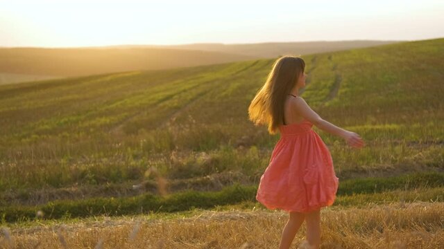 Happy long haired young woman in red summer dress spinning around on golden wheat field at sunset.