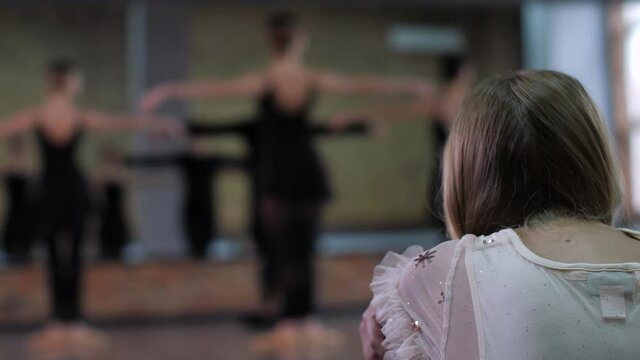 Rear view of blonde little girl quietly watching young female dancers practicing in ballroom blurry on background. Dreaming of being ballerina child enjoying dancer's lesson while sitting separately
