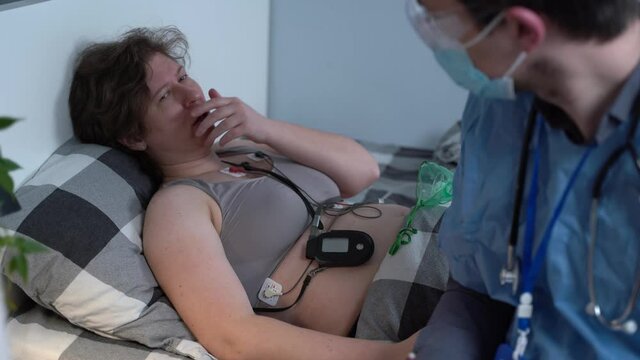 Covid 19, coronavirus. Doctor home visiting during quarantine, patient woman lying in bed with oxygen mask, medical worker doing electrocardiogram checking heart function. First aid, paramedicine