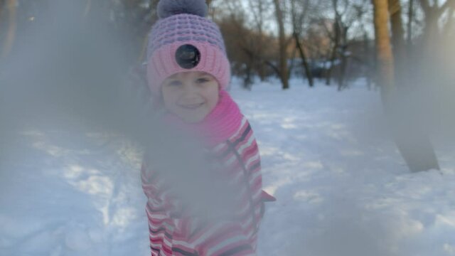 Cheerful child girl throwing snowball into camera in winter park. Portrait of funny children kid throw snowflakes, fooling around, smiling, outdoors at sunset. Traveling tourism, Christmas holidays