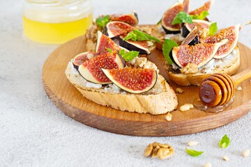 Crispy bruschetta (toast) with soft ricotta, ripe figs, walnuts and pine nuts, mint and honey on a light background. Figs fruit toast on a wooden board with honey and walnut.
