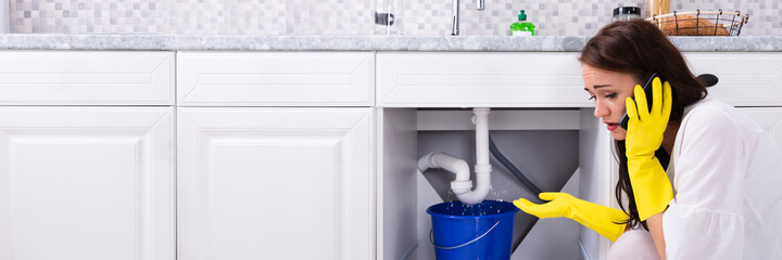 Woman Calling Plumber In Front Of Water Leaking From Pipe