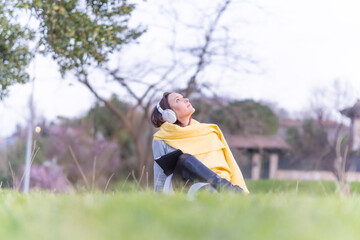 beautiful young woman in gray dress, yellow scarf and hat sitting in a park on the grass watching media on the tablet wearing headphones. relaxation lifestyle concept. businesswoman