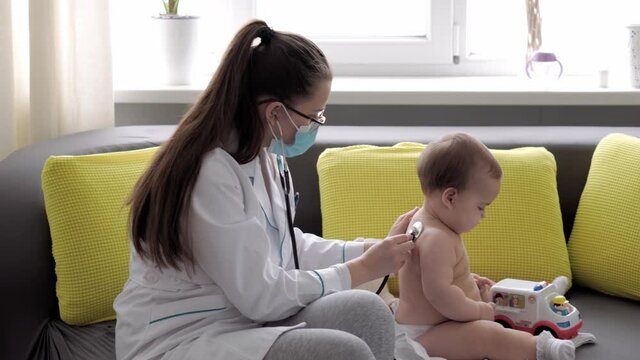 medicine and health, pediatrics, covid-19 concept - close-up young woman nurse or doctor pediatrician of Caucasian Slavic ethnicity examines infant 8-12 months baby on yellow gray sofa opposite window