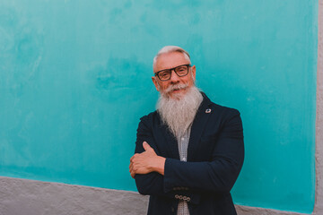 an older man in hipster clothes and glasses and a long white beard poses on a blue wall focus on...