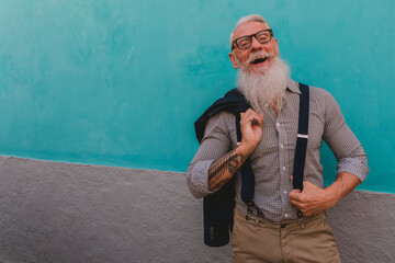 an older man in hipster clothes and glasses and a long white beard poses on a blue wall focus on...