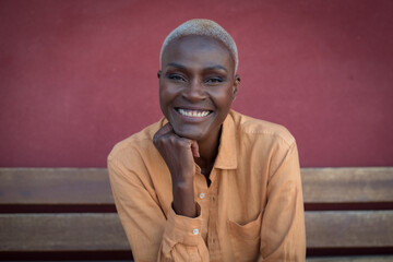 A black adult woman is sitting on a wooden bench and a red wall is in the background. She is relaxed. Focus on the face