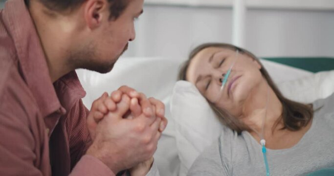 Close up of young man holding hand of sick mother sleeping in hospital bed
