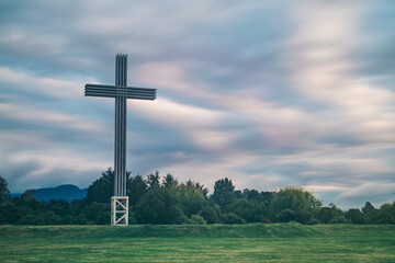 Huge cross in a park with some green trees, on a cloudy day and some hills in the background 