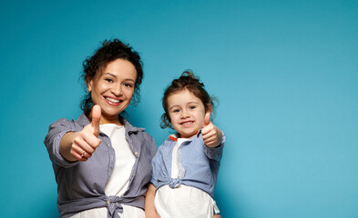 Beautiful mom and daughter equally dressed showing thumbs up and cute smiling while posing to camera on blue background with copy space