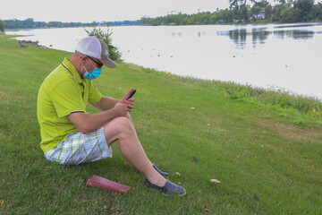 caucasian male wearing a face mask texting while sitting on the grass outside.