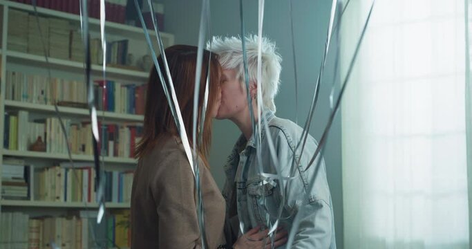 Cinematic shot of carefree happy homosexual female gay and transgender man couple in love enjoying time together is hugging and kissing with floating balloons to celebrate their forever love at home.