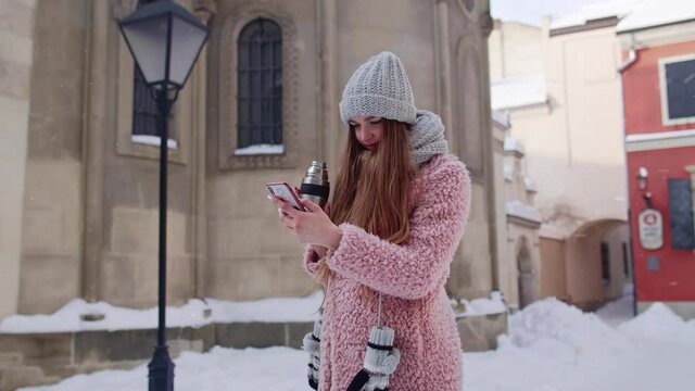 Traveler woman in city holding mobile phone, chatting with friends and family, drinking hot drink tea, coffee. Snowy weather. Girl blogger tourist publishing new photo picture post on social media