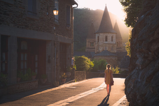 A young female tourist in a summer dress explores the quaint and charming medieval French village of Conques, Aveyron at sunset or sunrise in Occitanie France.