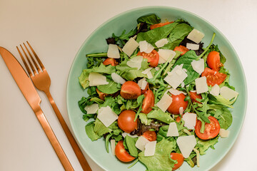 different salad leaves with cherry tomatoes and parmesan cheese in a green plate on a white background. Picture of salad for restaurant or pizzeria. Arugula, spinach, cherry tomatoes. Fork and knife