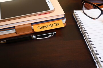 Corporate Tax has written on the notebook page label with a pen, smartphone, and eyeglasses on the notebook 