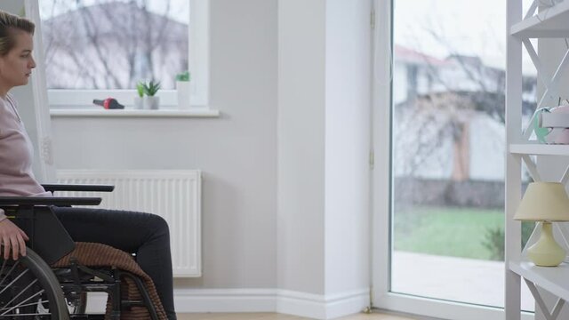Side view of young lonely disabled woman rolling wheelchair to glass door and looking outside. Sad Caucasian paralyzed invalid spending day at home alone. Hopelessness and disability concept.