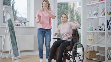 Wide shot of cheerful young woman dancing with handicapped friend indoors. Portrait of positive Caucasian paralyzed lady having fun with friend at home. Paralysis and happiness concept.