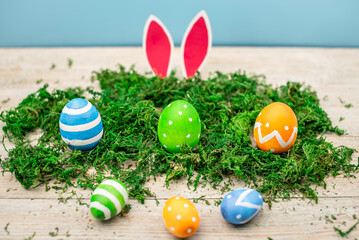 Bright colorful Easter eggs and rabbit ears on a blue and wooden background with green moss