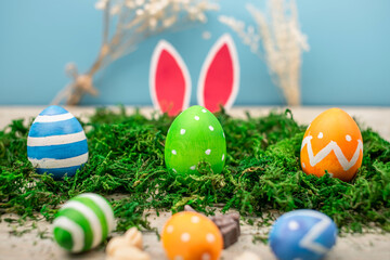 Bright colorful Easter eggs and rabbit ears on a blue and wooden background with green moss