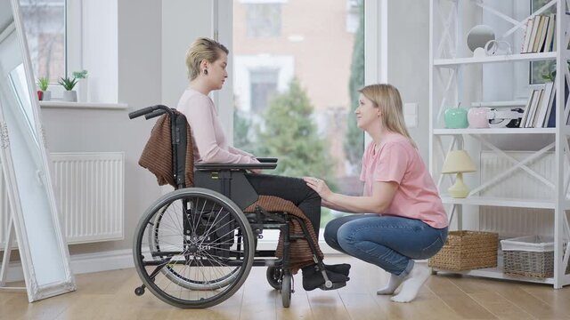 Side view wide shot of young paralyzed disabled woman on wheelchair talking with supportive friend at home. Upset paraplegic Caucasian invalid supported by friend indoors. Disability lifestyle.