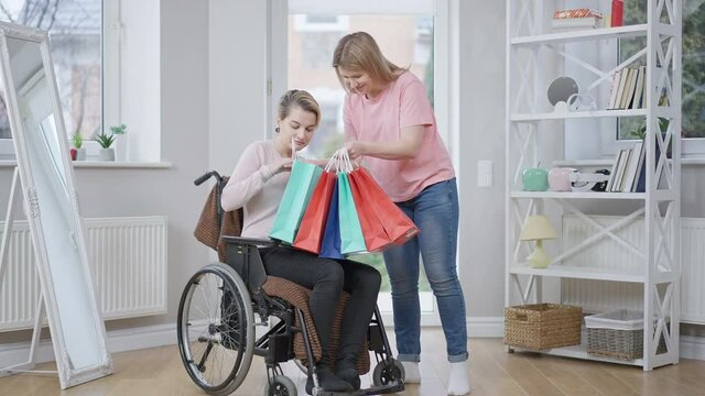 Wide shot portrait of smiling young woman bringing shopping bags for paraplegic friend. Positive Caucasian lady giving gifts to disabled beautiful invalid on wheelchair. Slow motion.