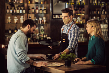 Happy waiter with face mask serving coffee to a couple in cafe.