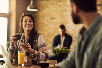 Happy woman using cell phone while talking to a friend and drinking coffee in a cafe.