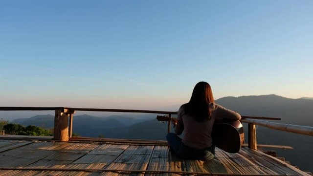 Rear view of a female sitting and playing guitar on wooden balcony with a beautiful mountain view before sunset