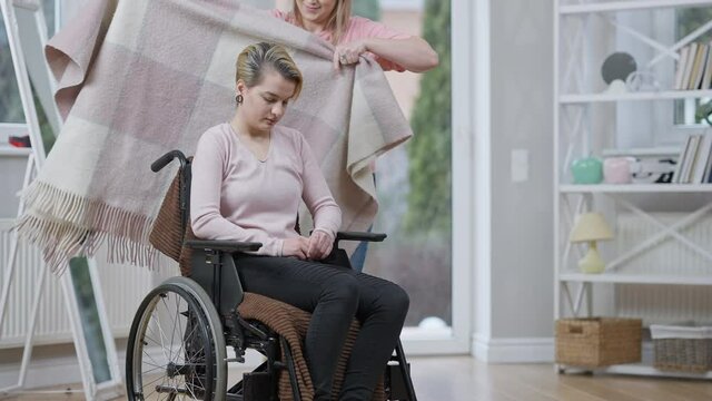 Positive young woman walking to disabled lady in wheelchair and covering friend with blanket. Stressed Caucasian paraplegic invalid smiling looking at caregiver. Lifestyle and disability.