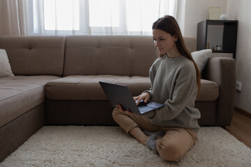 Fototapeta premium The girl sits on the floor next to the sofa, holding a laptop in her hands.