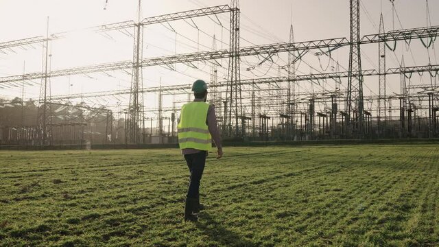 Electrical engineer wearing a helmet and safety vest walking near high voltage electrical lines towards power station during sunset shot in 4k super slow motion