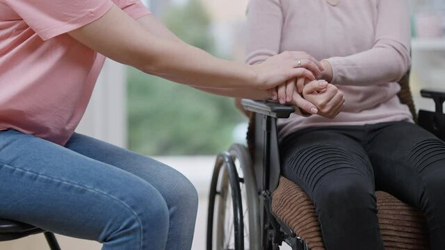 Unrecognizable young paralyzed woman and friend holding hands sitting indoors. Slim Caucasian empathic lady cheering up depressed paralyzed friend at home. Support and handicap.