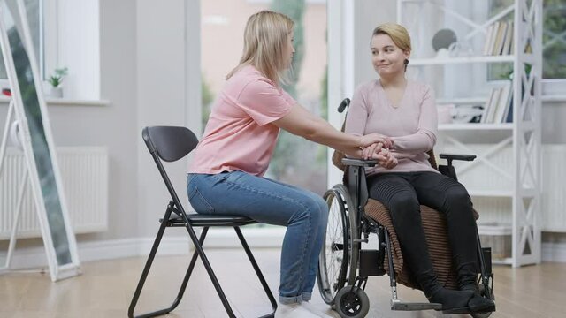 Wide shot of sad disabled young woman in wheelchair and supportive friend holding hands. Blond Caucasian empathic lady taking care of paraplegic invalid after trauma. Support and friendship.