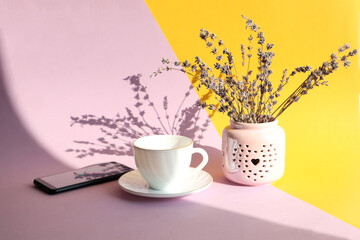A bouquet of lavender with a cup of tea and a phone on a yellow-pastel background in the sun with a shadow from objects, side view
