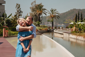 Fototapeta na wymiar Loving couple enjoying honeymoon in luxury hotel, walking through grounds with palm trees and swimming pool. Happy lovers on romantic trip have fun on summer vacation. Concept romance and relaxation
