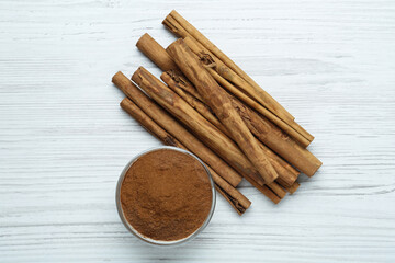 Aromatic cinnamon powder in bowl near sticks on white wooden table, flat lay