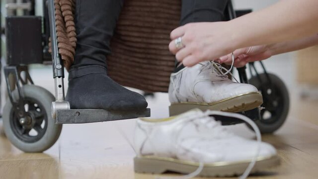 Female hands tying shoelaces on shoes of unrecognizable paraplegic person in wheelchair. Young Caucasian volunteer taking care of disabled woman. Friend helping paralyzed woman at home.