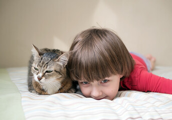A Caucasian boy in casual wear with a cat lie on the bed and look at the camera. Portrait. Close-up