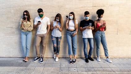 group of multiracial friends with face masks in front of a clean wall using smartphones to sharing content on social networks. Covid-19 and technology lifestyle  concept - obrazy, fototapety, plakaty