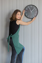 asian female young adult wearing black sleeveless shirt with green striped apron stand smiling happily holding and adjusting black round metal clock with white numbers and hands on stripe wall