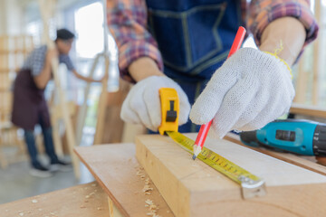 Worker hands wearing white gloves holding red pencil and yellow measure tape measuring the wood board on the table while male colleague choosing the wooden sticks in blurred background