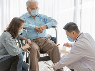 A young asian male doctor wearing white lab coat and face mask checking left knee of old fat gray hair patient wearing light blue shirt and brown pants while he sitting on wheelchair 