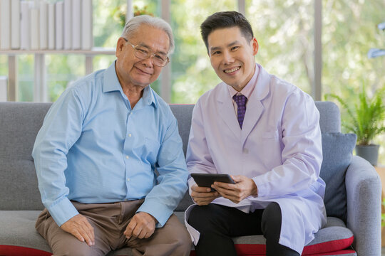 An Old Fat Asian Male Patient With White Gray Hair Wearing Light Blue Shirt With Brown Pants And A Young Handsome Doctor Wearing White Lab Coat Hold Small Black Tablet In His Hand 