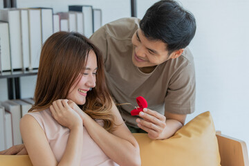 A young asian black hair male wearing brown shirt showing red diamond ring box in the left hand trying to propose marriage when asian long brown hair female smile exciting and sitting on orange sofa 