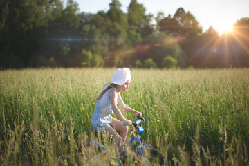 A six-year-old girl rides a bicycle in the field. A large white hat on her head. Sunset.