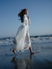 Fototapeta na wymiar A woman in a white dress walks on the wet sand on the shore of the ocean in full growth